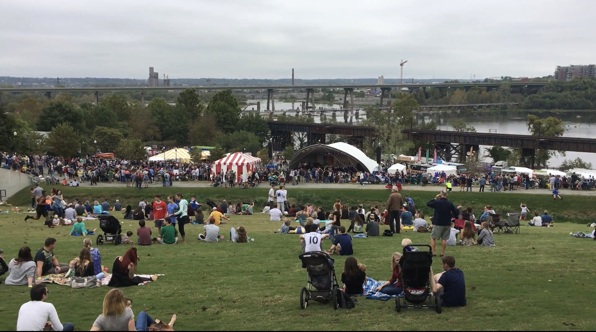 A crowd of people walking musicians perform on stage at the Richmond Folk Festival near parking and the James River waterfront.